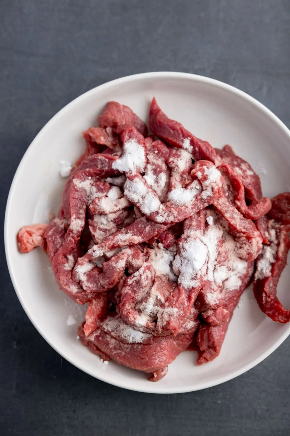 Overhead image of a white bowl on a gray surface filled with strips of raw beef steaks coated in baking soda.