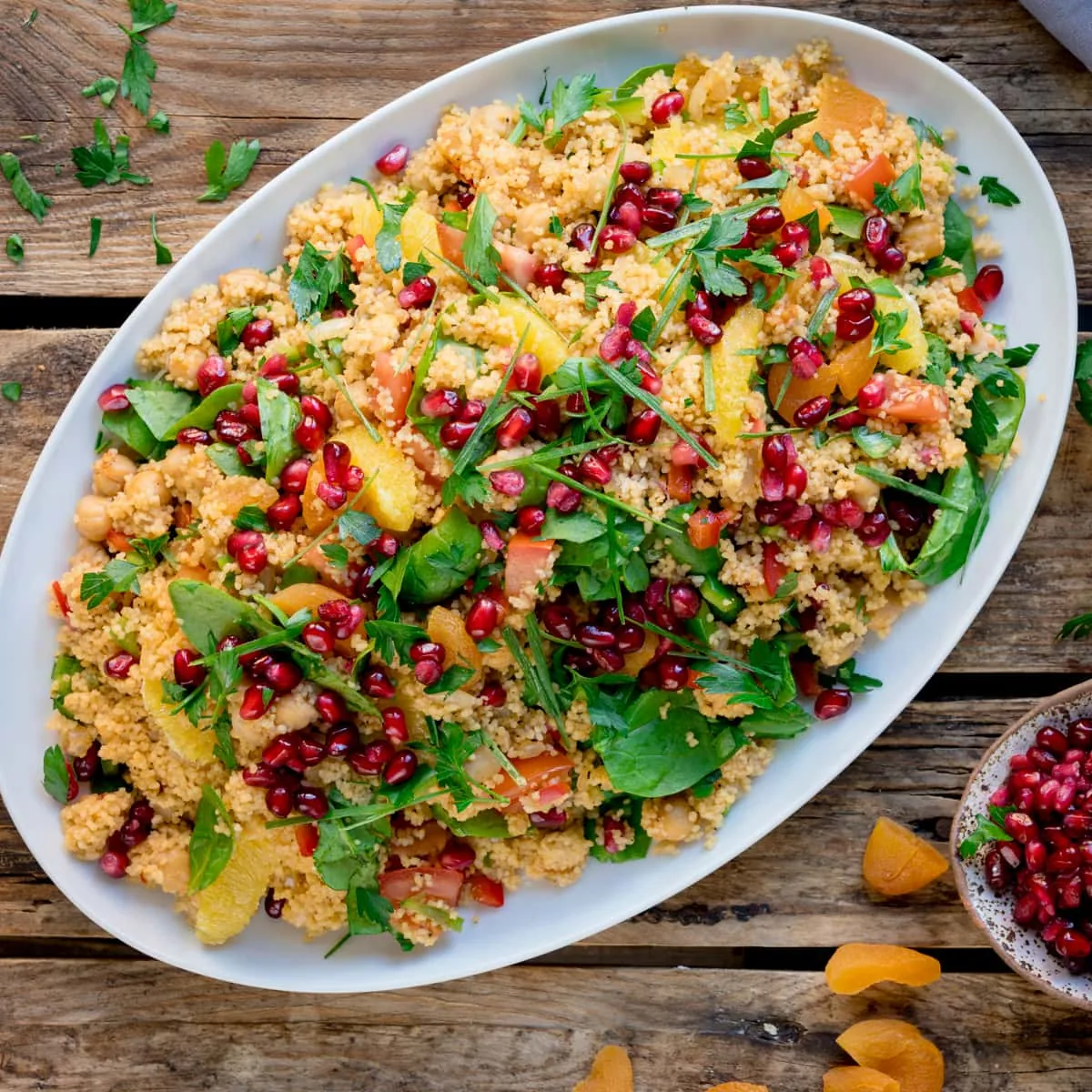 Overhead square image of a large oval white plate filled with Moroccan style couscous with oranges and pomegranate. The plate is on a wooden table, next to a dish of pomegranate. There are chopped dried apricots and fresh herbs scattered around.