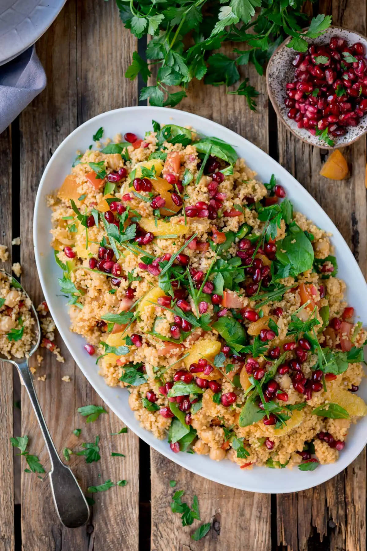 Overhead image of a large oval white plate filled with Moroccan style couscous with oranges and pomegranate. The plate is on a wooden table, next to a spoon filled with couscous, a dish of pomegranate and fresh herbs scattered around.