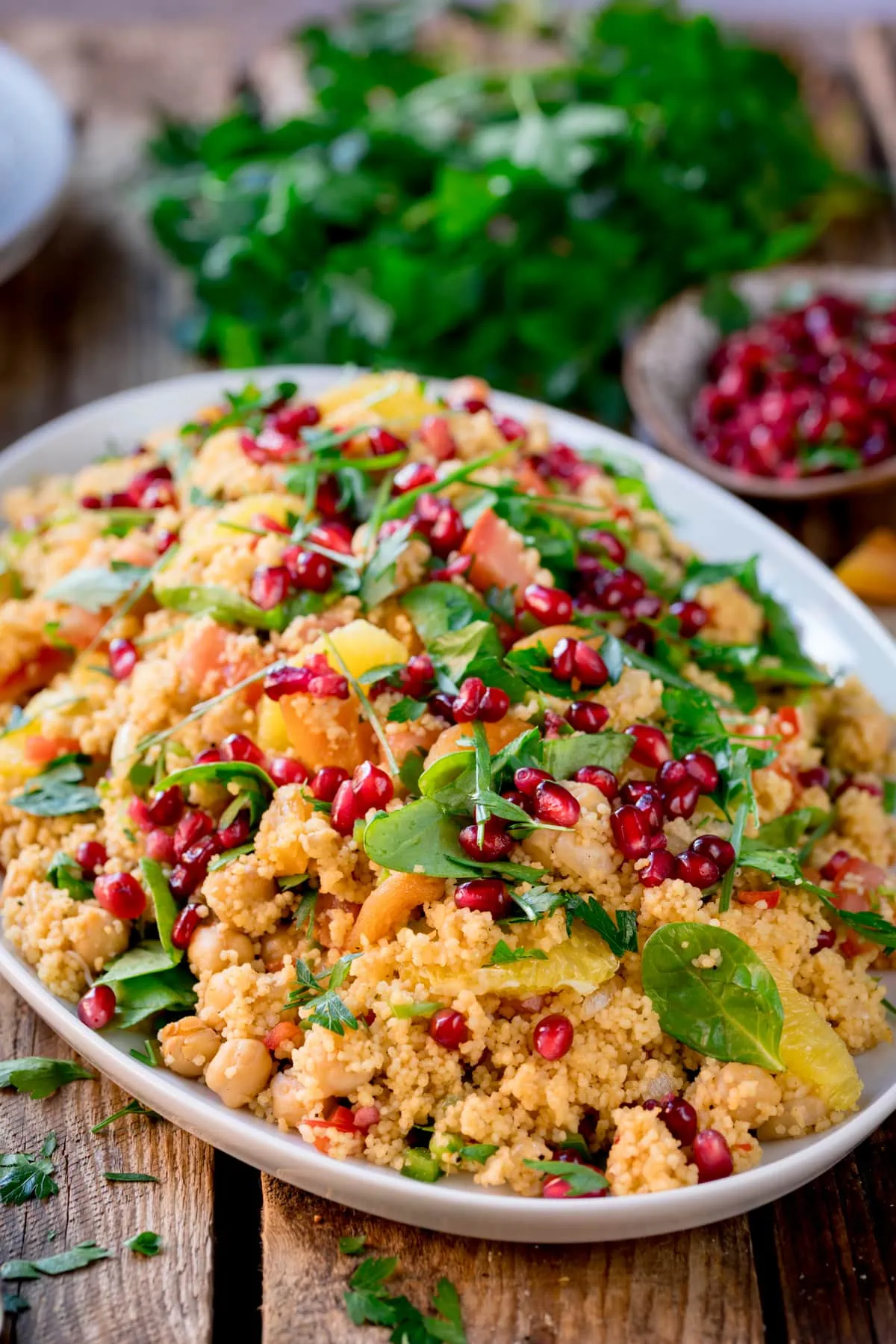 Side-on image of a large oval serving plate filled with Moroccan style couscous, topped with herbs and pomegranate. The plate is on a wooden table and there are fresh herbs and a little bowl of pomegranate in the background.