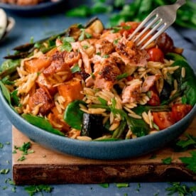 A blue bowl full of orzo pasta salad with salmon on a wooden chopping board.