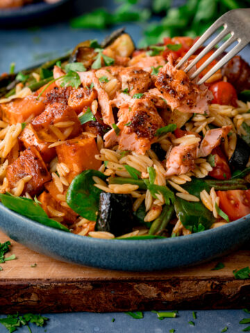 A blue bowl full of orzo pasta salad with salmon on a wooden chopping board.