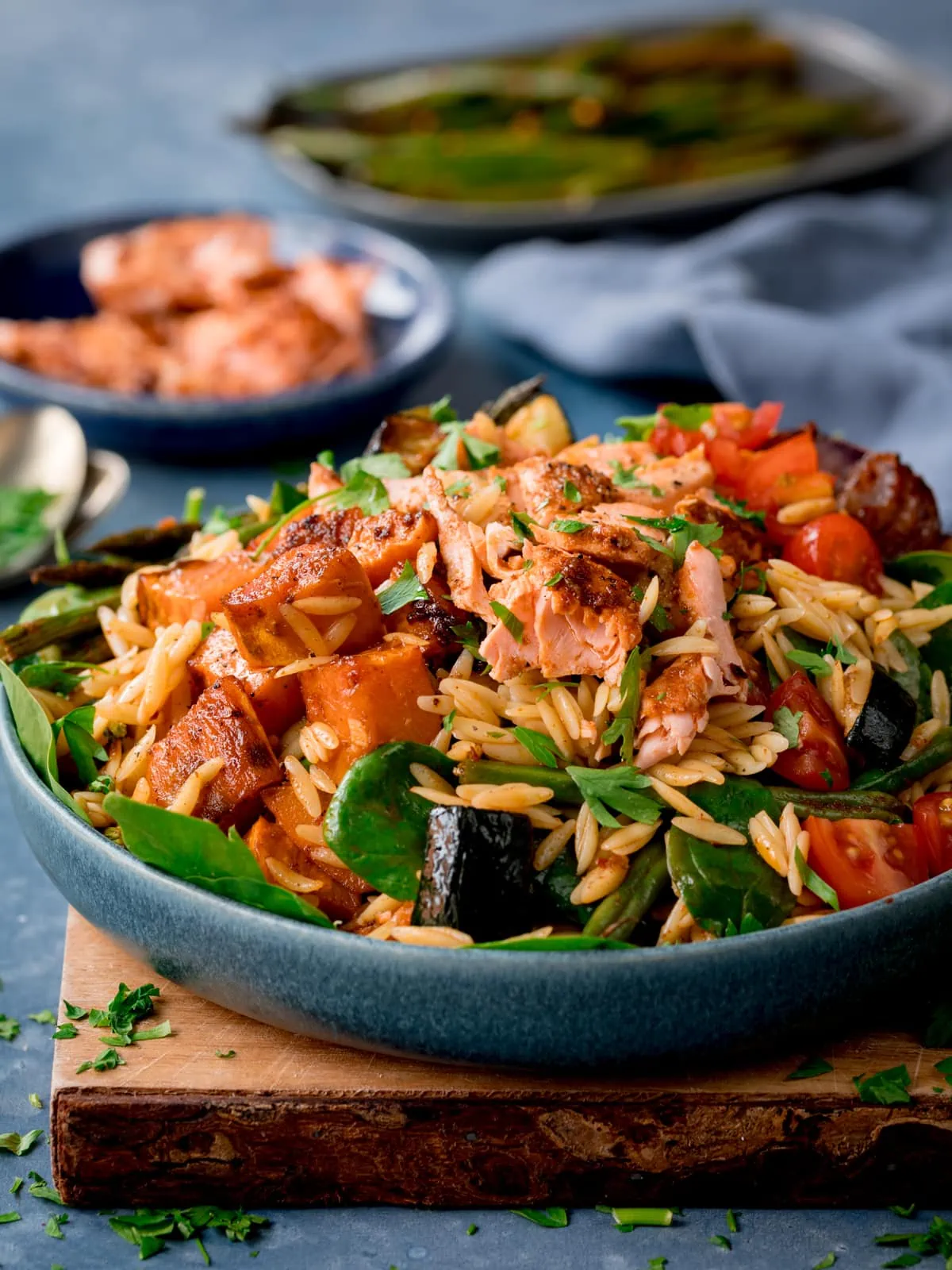 Side photo of orzo pasta salad with salmon in a blue bowl sat on a wooden board.