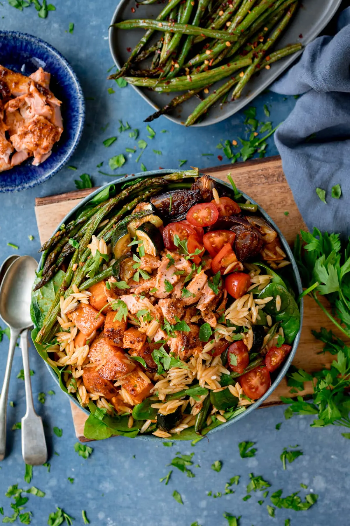 A bowl of orzo pasta salad with salmon and roasted vegetables on a wooden board with a blue background.