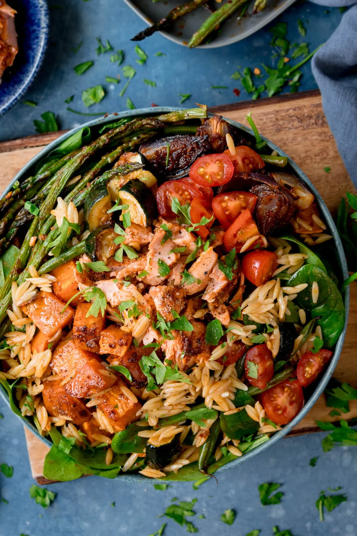 A bowl full of orzo pasta salad with salmon and roasted vegetables on a wooden board with a blue background.