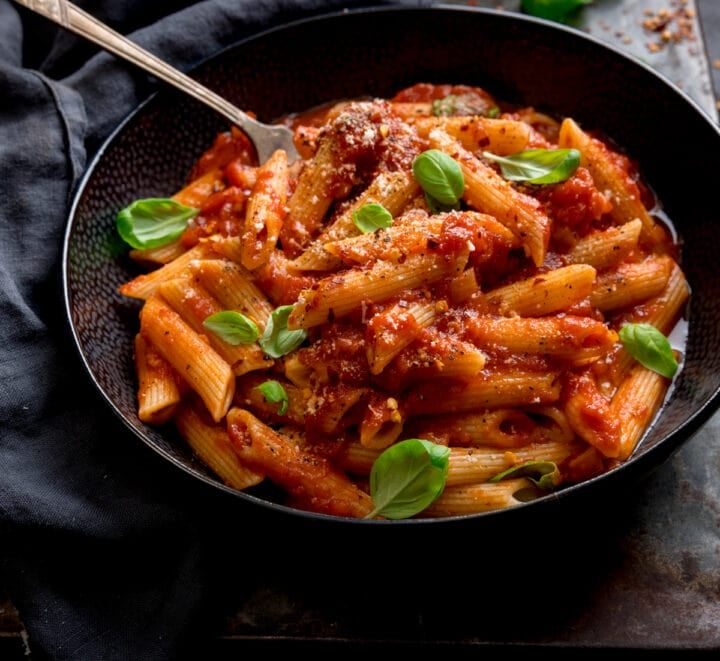 A black bowl filled with penne arrabiata, with fresh baby basil leaves sprinkled on top. There is a fork sticking out of the bowl. The bowl is in a dark surface, next to a grey napkin.