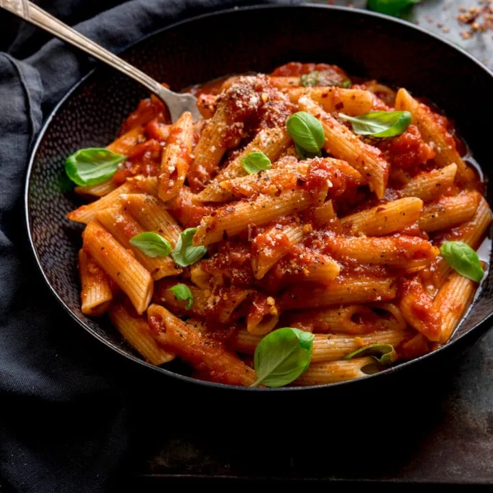 A black bowl filled with penne arrabiata, with fresh baby basil leaves sprinkled on top. There is a fork sticking out of the bowl. The bowl is in a dark surface, next to a grey napkin.