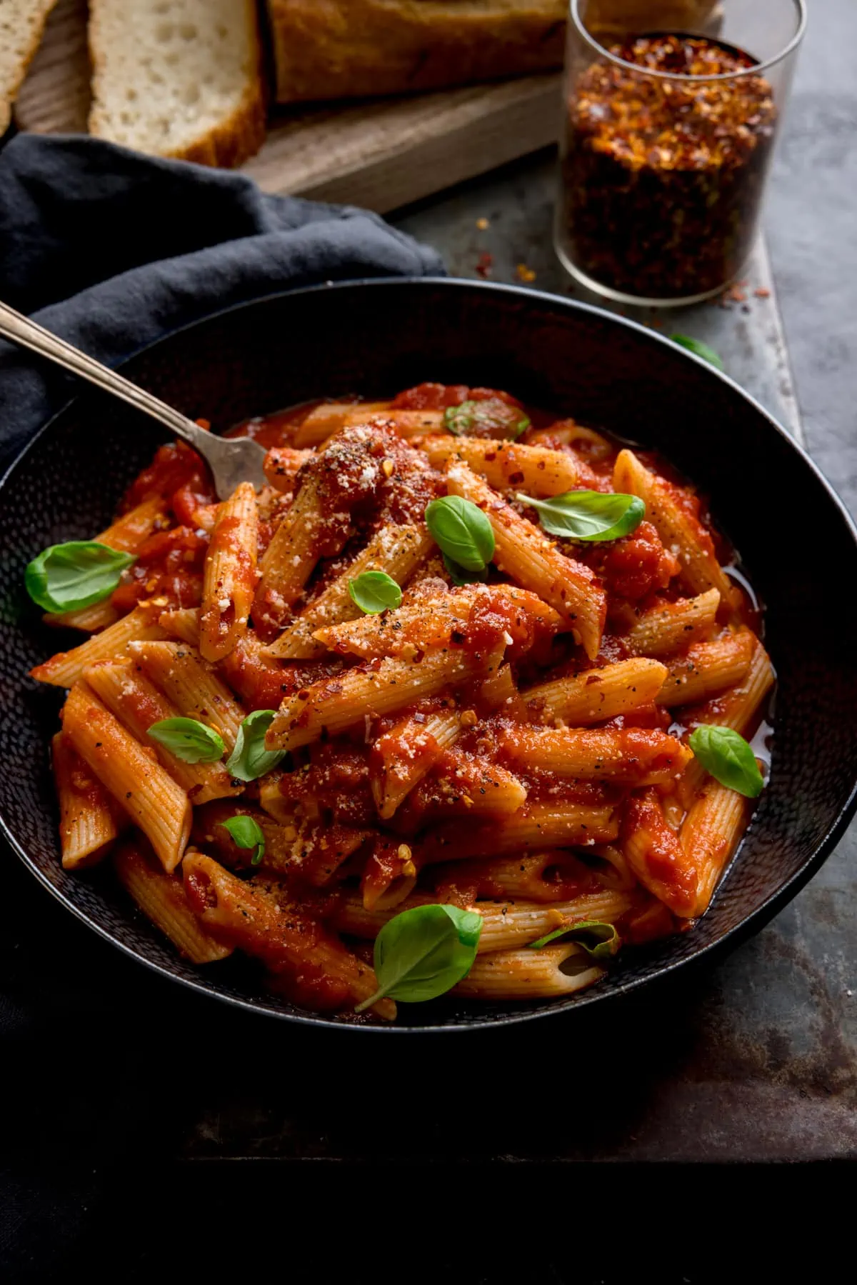 A black bowl filled with penne arrabiata, with fresh baby basil leaves sprinkled on top. There is a fork sticking out of the bowl. The bowl is in a dark surface and there is a loaf of bread with a couple of sliced pieces and a jar of chilli flakes at the top of the frame.