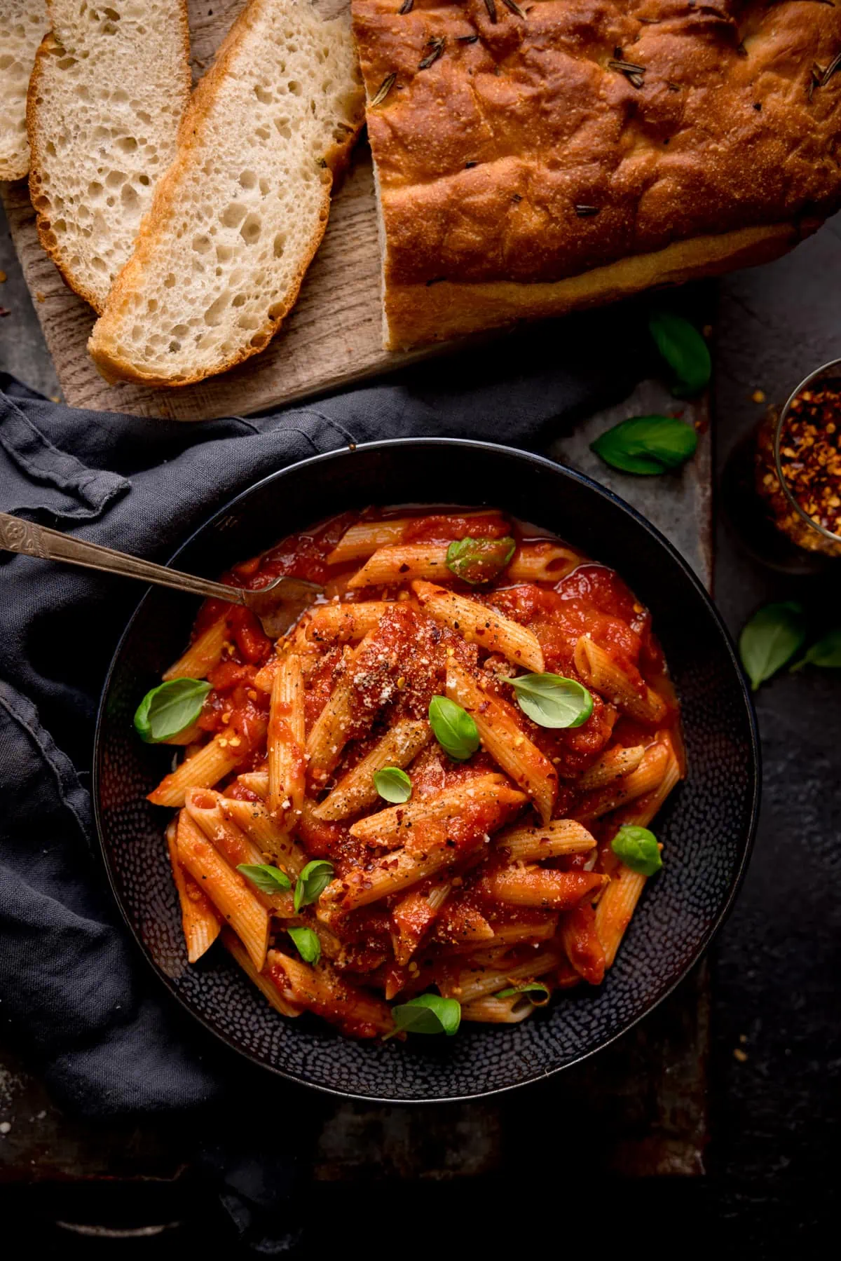 Overhead image of a black bowl filled with penne arrabiata, with fresh baby basil leaves sprinkled on top. There is a fork sticking out of the bowl. The bowl is in a dark surface and there is a loaf of bread with a couple of sliced pieces at the top of the frame.