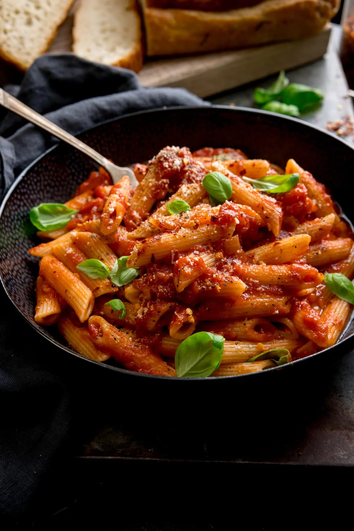 A black bowl filled with penne arrabiata, with fresh baby basil leaves sprinkled on top. There is a fork sticking out of the bowl. The bowl is in a dark surface and there is a loaf of bread with a couple of sliced pieces at the top of the frame.