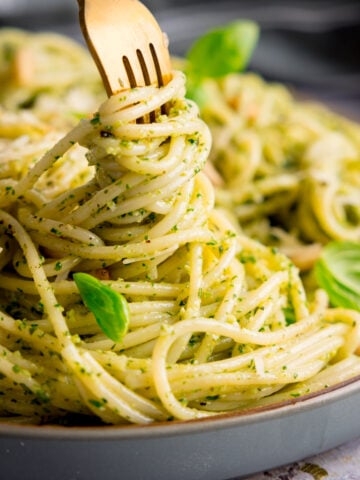Square image of pesto spaghetti on a light grey plate being swirled with a gold fork. The pasta is sprinkled with baby basil leaves, toasted pine nuts and parmesan. The plate is on a light grey surface.