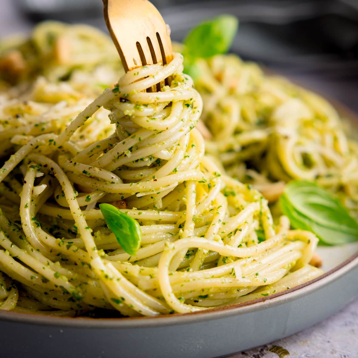 Square image of pesto spaghetti on a light grey plate being swirled with a gold fork. The pasta is sprinkled with baby basil leaves, toasted pine nuts and parmesan. The plate is on a light grey surface.