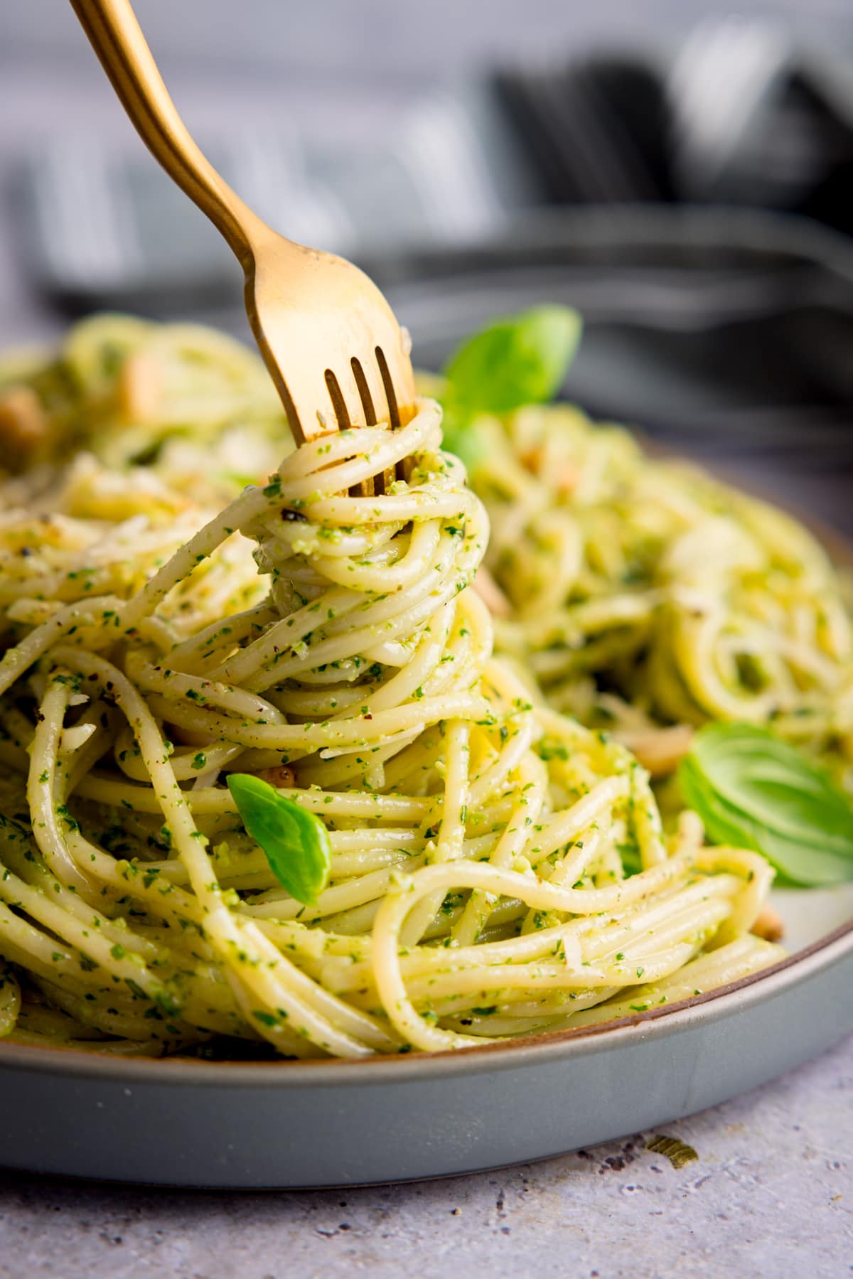 Pesto spaghetti on a light grey plate being swirled with a gold fork. The pasta is sprinkled with baby basil leaves, toasted pine nuts and parmesan. The plate is on a light grey surface.