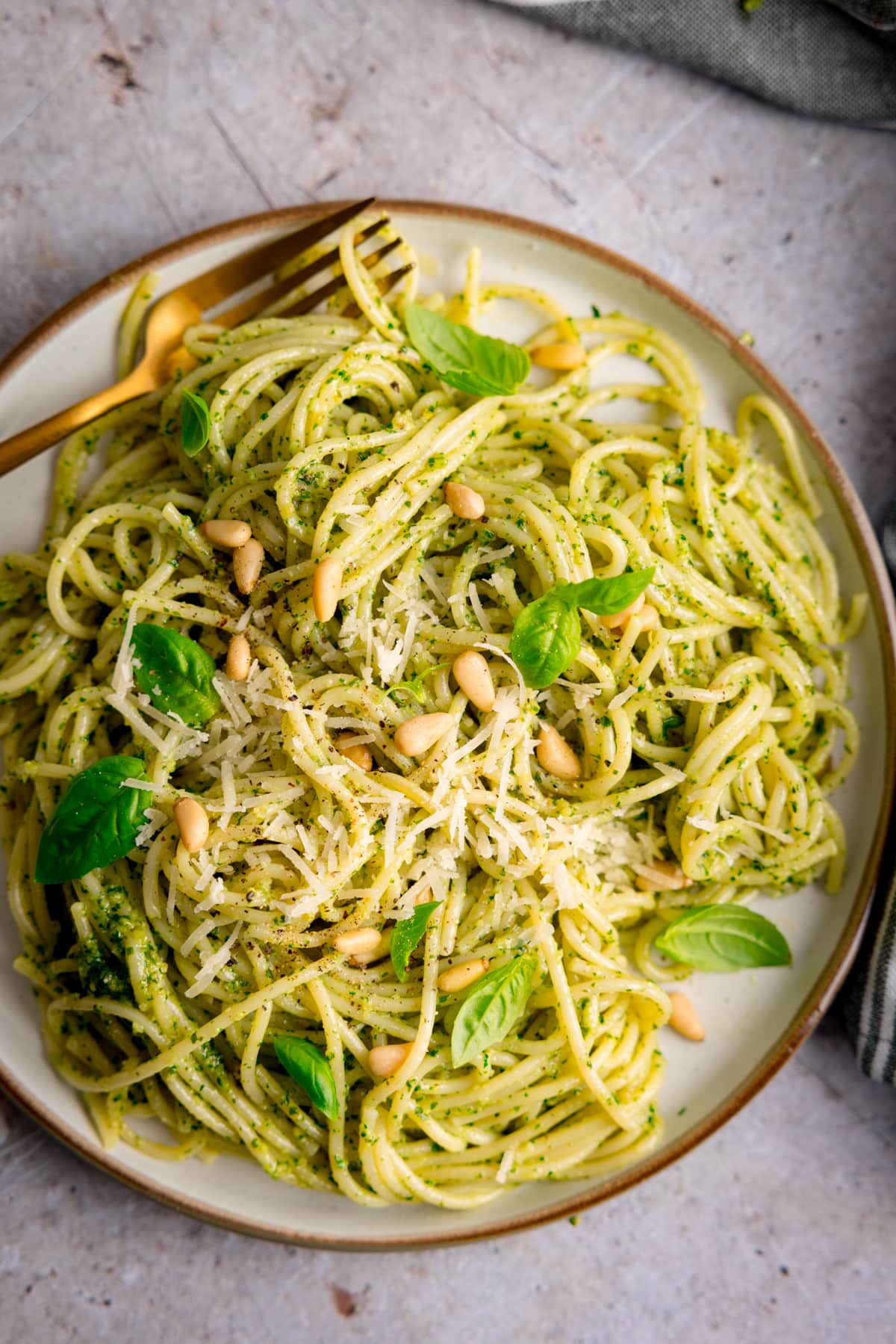 Overhead image of pesto spaghetti on a light grey plate. The pasta is sprinkled with baby basil leaves, toasted pine nuts and parmesan. The plate is on a light grey surface and there is a gold fork on the plate.