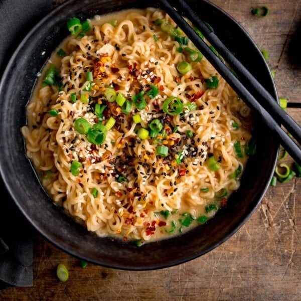 Overhead square image of a black bowl filled with ramen noodles topped with chilli flakes, spring onions and mixed sesame seeds. There is a pair of black chopsticks resting on top of the bowl. The bowl is on a wooden table.