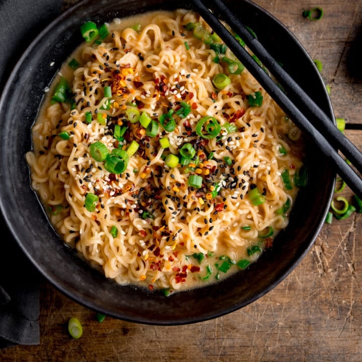 Overhead square image of a black bowl filled with ramen noodles topped with chilli flakes, spring onions and mixed sesame seeds. There is a pair of black chopsticks resting on top of the bowl. The bowl is on a wooden table.