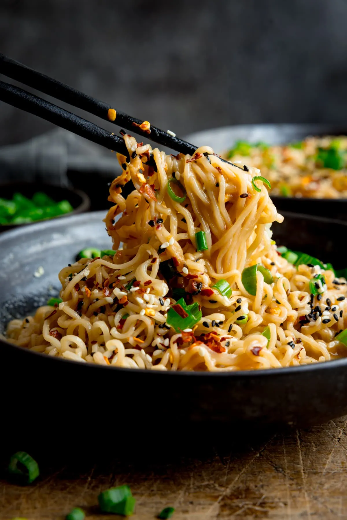 Side-on image of a black bowl, filled with ramen noodles. A pair of black chopsticks are being used to lift the noodles. The bowl is on a wooden table. There are spring onions scattered around and a further bowl of noodles in the background.