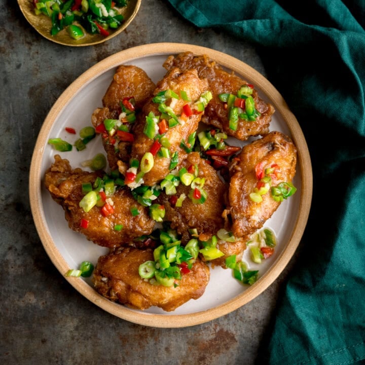 Square overhead image of crispy salt and pepper chicken wings on a white plate on a rustic metal surface. There is a green napkin and a small gold dish with some of the chilli-salt-pepper topping in it.