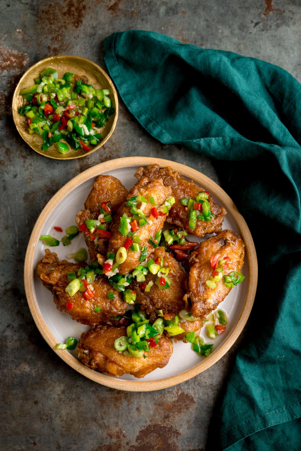 Overhead image of crispy salt and pepper chicken wings on a white plate on a rustic metal surface. There is a green napkin and a small gold dish with some of the chilli-salt-pepper topping in it.
