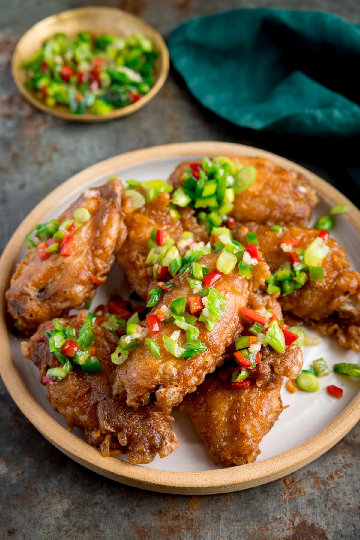 Tall side-on image of crispy salt and pepper chicken wings on a white plate on a rustic metal surface. There is a green napkin and a small gold dish with some of the chilli-salt-pepper topping in it at the top of the frame.