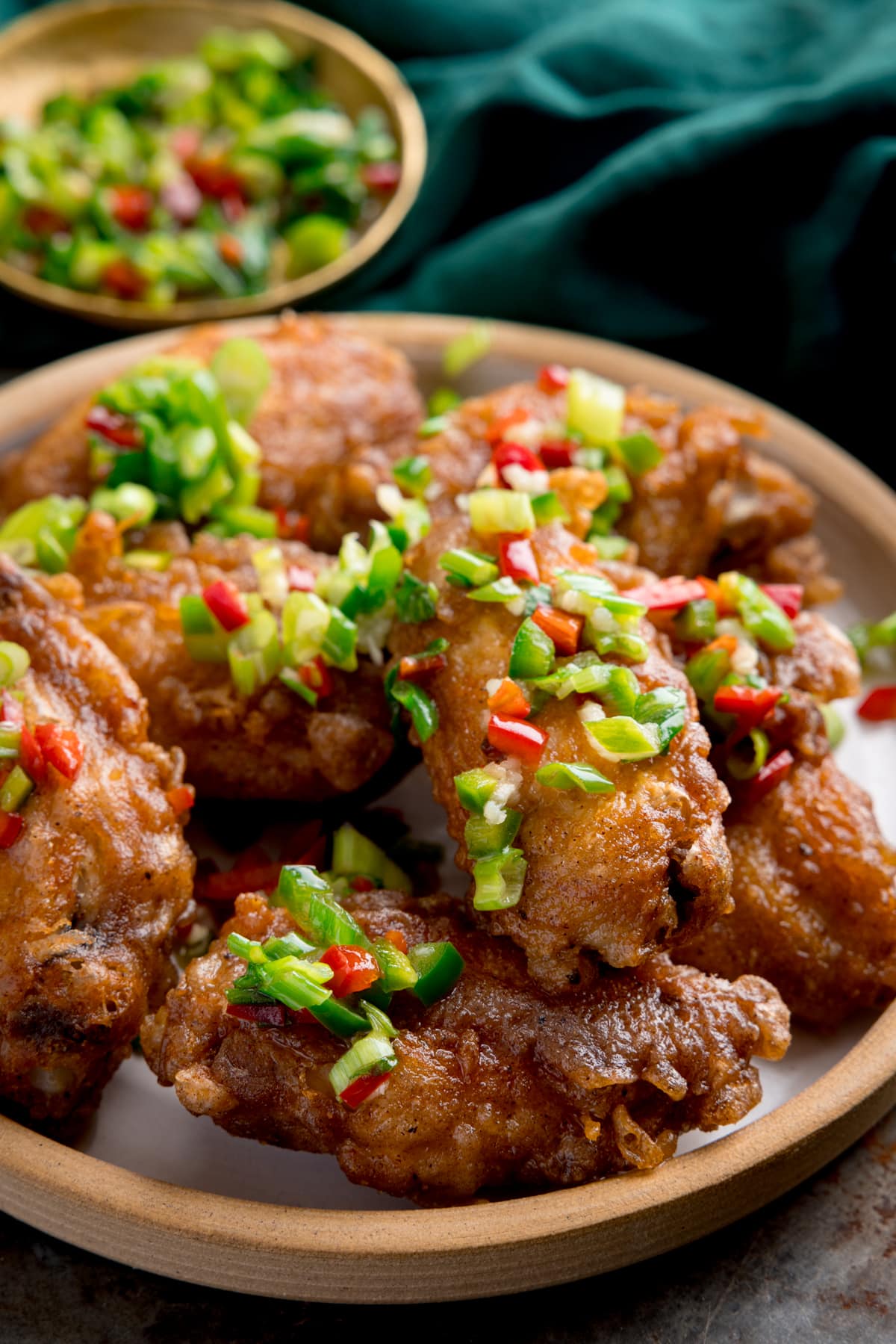 Tall side-on image of crispy salt and pepper chicken wings on a white plate on a rustic metal surface. There is a green napkin and a small gold dish with some of the chilli-salt-pepper topping in it at the top of the frame.