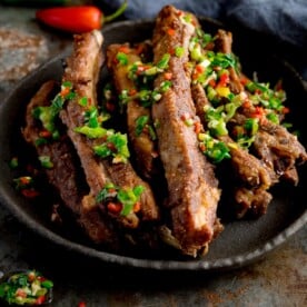 Square image of salt and pepper pork ribs on a black plate. The ribs are topped with chopped chillies and spring onions. The plate is on a dark background. There is a red chilli pepper and blue napkin at the top of the frame.