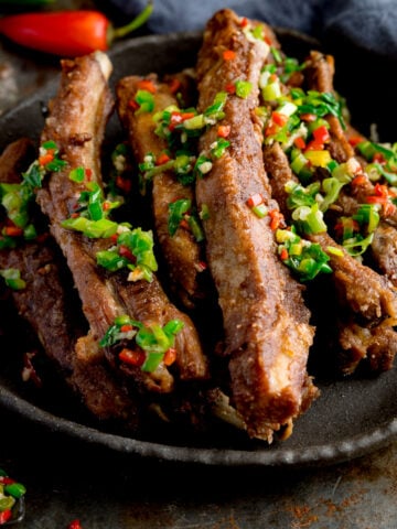 Square image of salt and pepper pork ribs on a black plate. The ribs are topped with chopped chillies and spring onions. The plate is on a dark background. There is a red chilli pepper and blue napkin at the top of the frame.