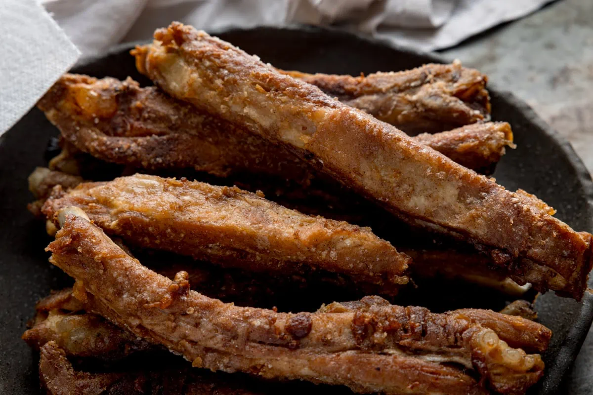 Wide image of crispy pork ribs on a dark plate next to a beige napkin.