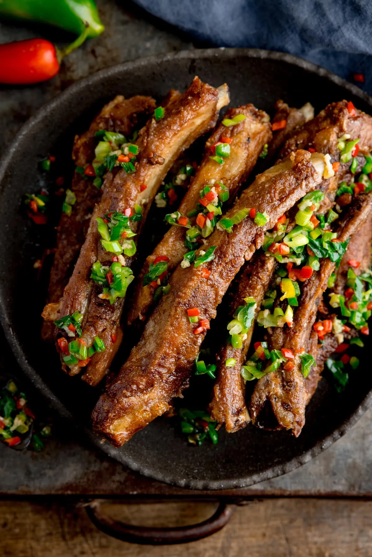 Overhead image of salt and pepper pork ribs on a black plate. The ribs are topped with chopped chillies and spring onions. The plate is on a dark background. There is a blue napkin and a red and green chilli pepper in the top of the frame.