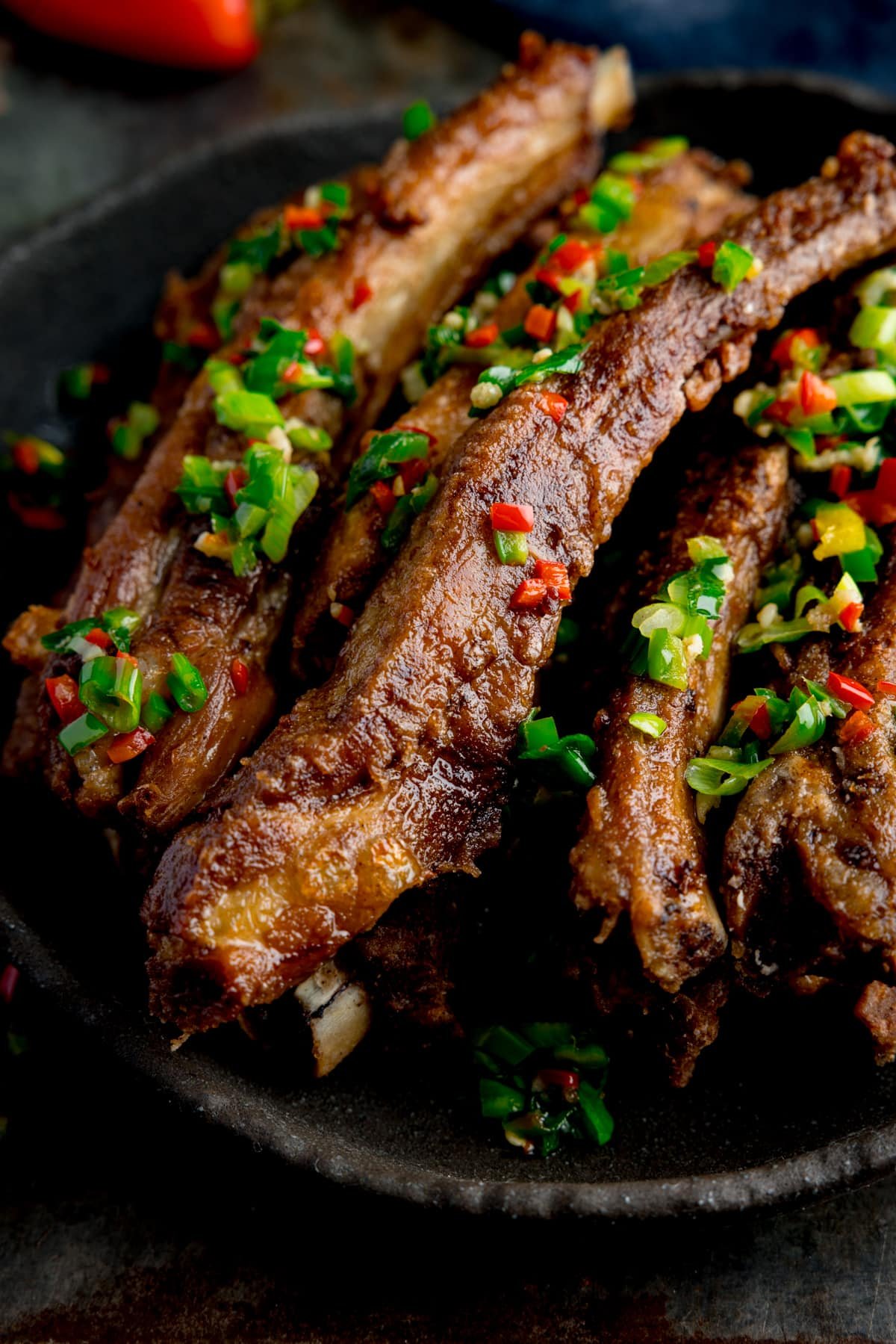 Salt and pepper the pork ribs on a black plate. The ribs are topped with chopped chillies and spring onions. The plate is on a dark background. There is a red chili pepper at the top of the frame.