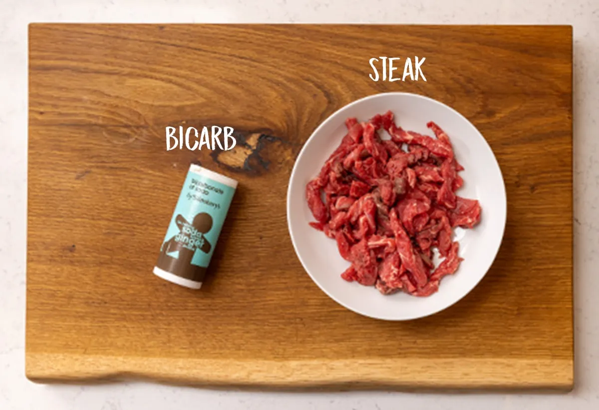 A white bowl filled with steak strips and a tub of bicarbonate of soda on a wooden board.