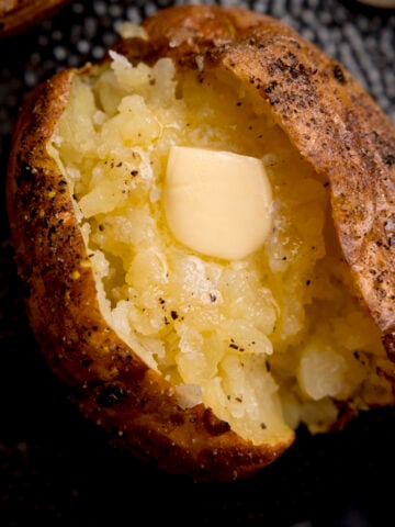 Overhead close-up image of a seasoned baked potato, sliced down the middle. The inside of the potato has been fluffed and there's a knob of melting butter inside. The potato is on a black textured plate.