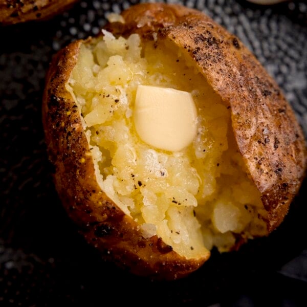 Overhead close-up image of a seasoned baked potato, sliced down the middle. The inside of the potato has been fluffed and there's a knob of melting butter inside. The potato is on a black textured plate.