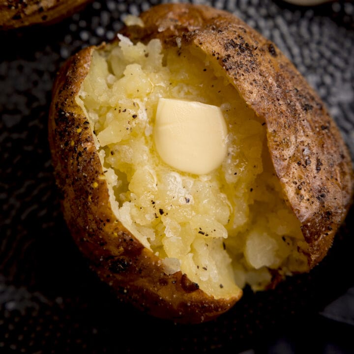 Overhead close-up image of a seasoned baked potato, sliced down the middle. The inside of the potato has been fluffed and there's a knob of melting butter inside. The potato is on a black textured plate.