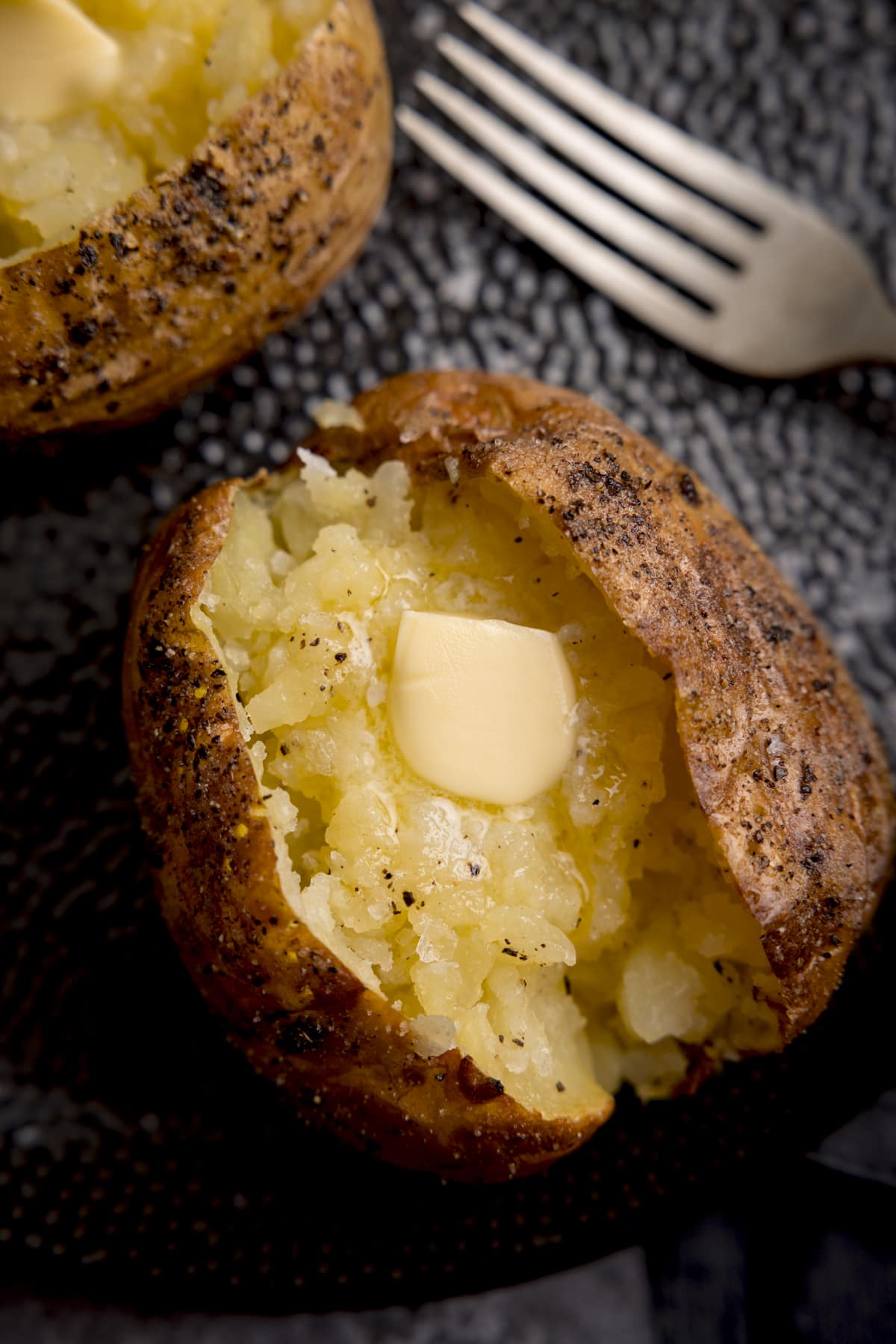 Overhead close-up image of a seasoned baked potato, sliced down the middle. The inside of the potato has been fluffed and there's a knob of melting butter inside. The potato is on a black textured plate. There is a silver fork just in view at the top right of the frame and a second baked potato just in view at the top left of the frame.