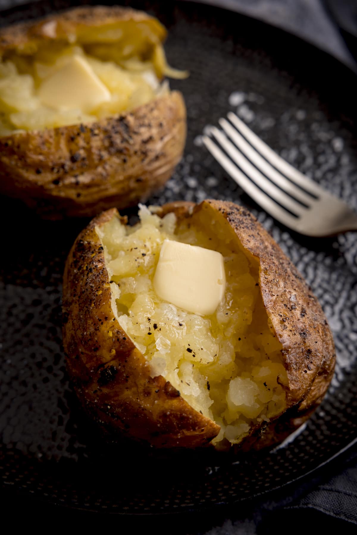 A seasoned baked potato, sliced down the middle on a black textured plate. The inside of the potato has been fluffed and there's a knob of melting butter inside. There is a silver fork just in view on the right of the frame and a second baked potato just in view at the top left of the frame.