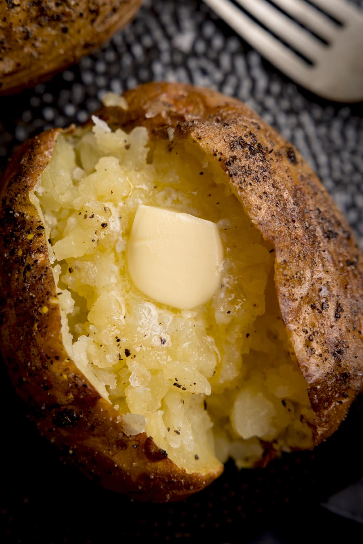 Overhead close-up image of a seasoned baked potato, sliced down the middle. The inside of the potato has been fluffed and there's a knob of melting butter inside. The potato is on a black textured plate. There is a silver fork just in view at the top right of the frame.