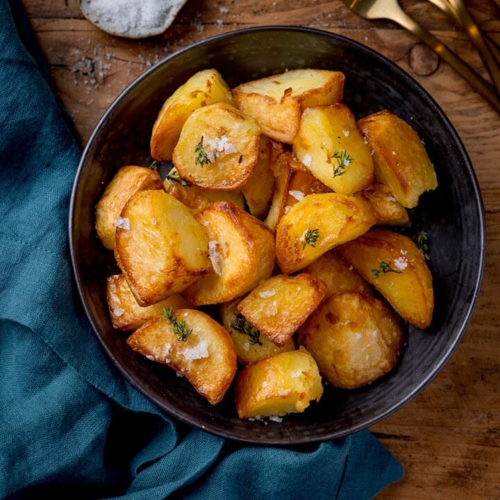 Square overhead image of roast potatoes piled up in dark bowl. The potatoes are sprinkled with thyme and salt. The bowl is on a wooden table next to a green napkin. There is a small dish of salt and gold cutlery partly in shot at the top of the frame.