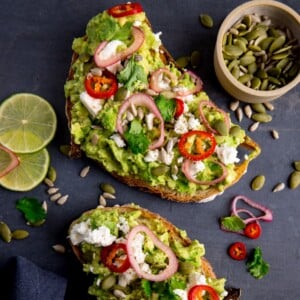 Two pieces of loaded avocado toast with feta, shallot, sliced red chilli, seeds and coriander (cilantro) on a dark grey background. There are ingredients scattered around and a pinch pot of seeds in the top right corner.