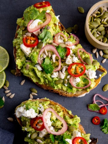 Two pieces of loaded avocado toast with feta, shallot, sliced red chilli, seeds and coriander (cilantro) on a dark grey background. There are ingredients scattered around and a pinch pot of seeds in the top right corner.