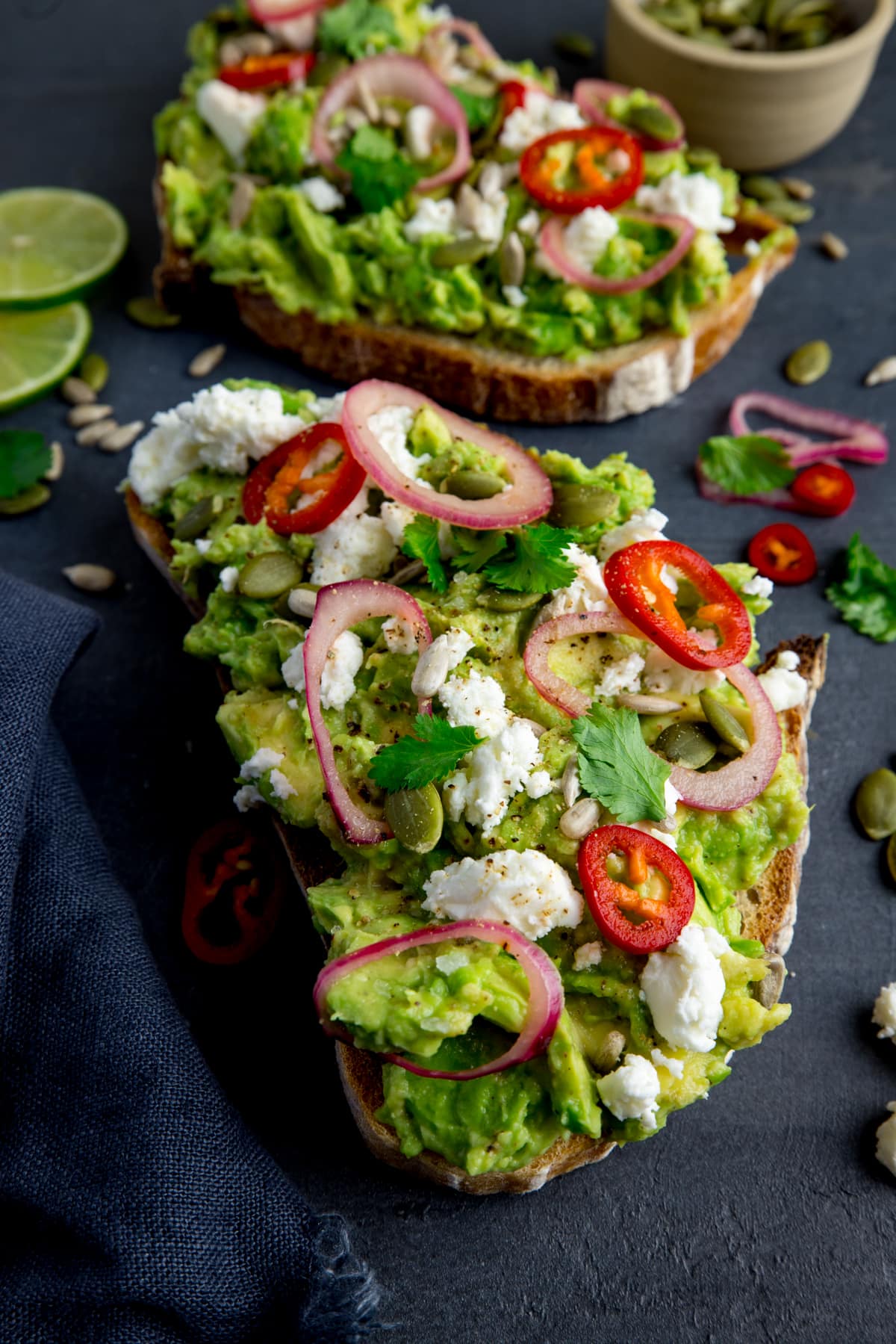 Two pieces of loaded avocado toast with feta, shallot, sliced red chilli, seeds and coriander (cilantro) on a dark grey background. There are ingredients scattered around and a dark grey napkin on the left of the frame. There is a pinch pot of seeds at the top right of the frame.