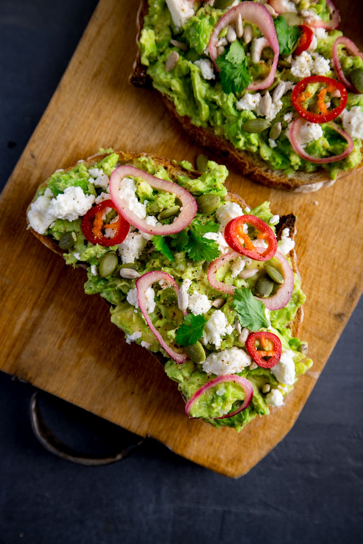 Two pieces of loaded avocado toast with feta, shallot, sliced red chilli, seeds and coriander (cilantro) on a wooden board on a dark grey background.