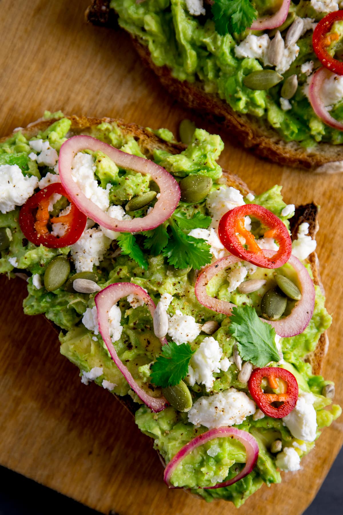 A piece of loaded avocado toast with feta, shallot, sliced red chilli, seeds and coriander (cilantro) on a wooden board. There is a further piece, just in shot at the top of the frame.