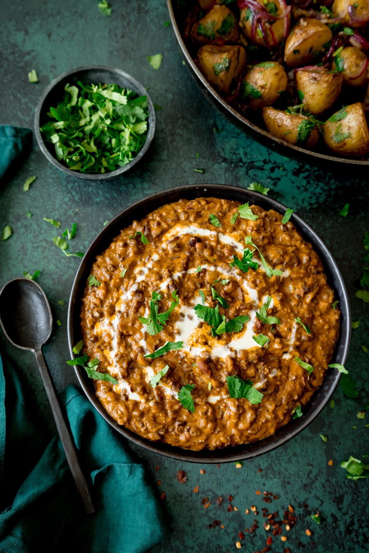 Black Lentil Dal in a dark bowl on a green board with coriander and potatoes in the background.