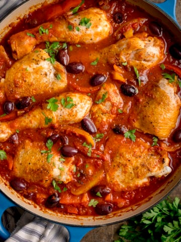 Overhead square image of chicken cacciatore in a large, shallow blue casserole dish. The dish is on a wooden table, next to some fresh herbs.