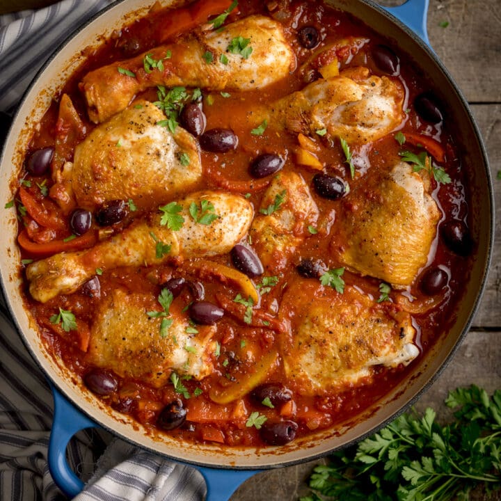 Overhead square image of chicken cacciatore in a large, shallow blue casserole dish. The dish is on a wooden table, next to some fresh herbs.