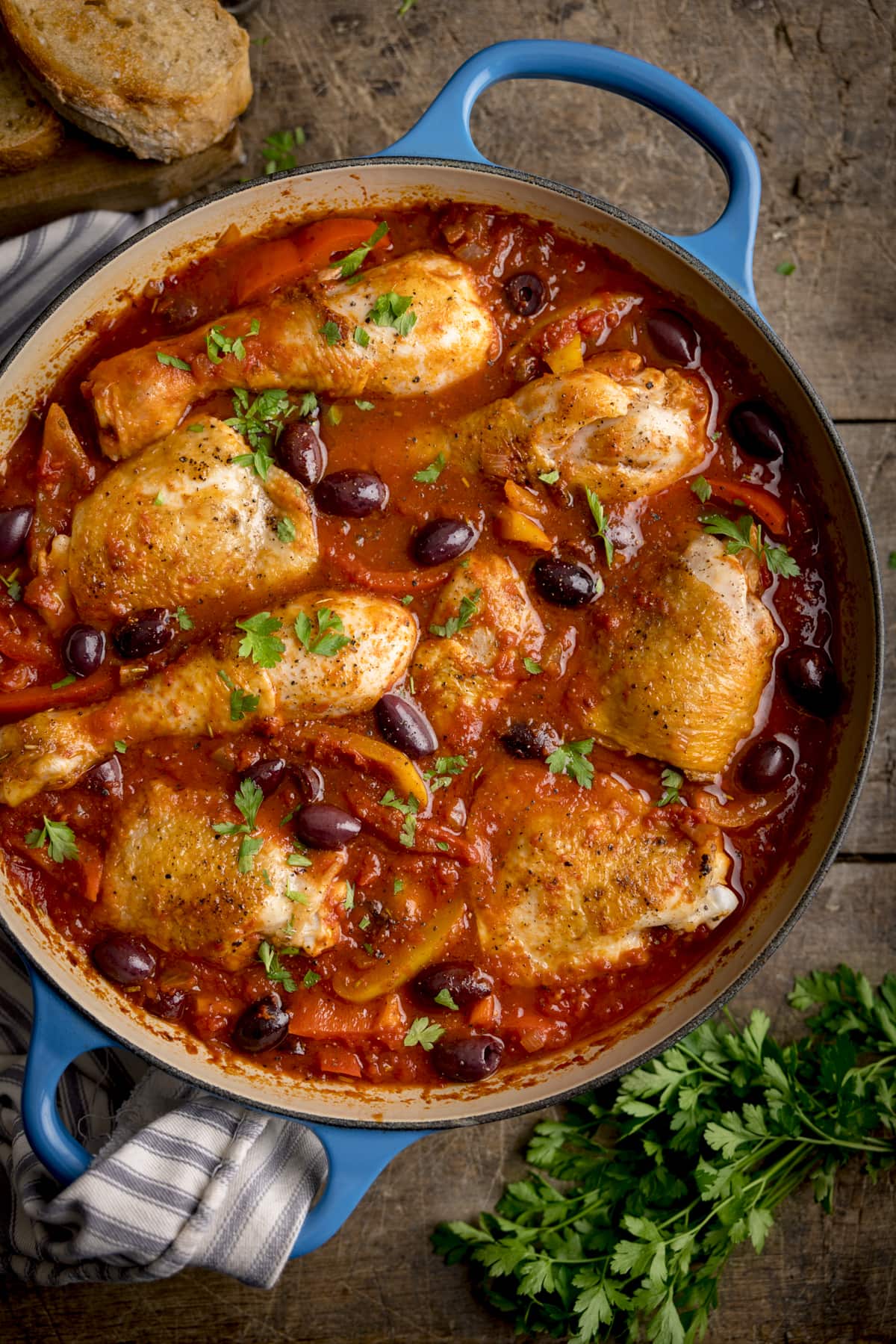Overhead image of chicken cacciatore in a large, shallow blue casserole dish. The dish is on a wooden table, next to some fresh herbs and sliced bread.