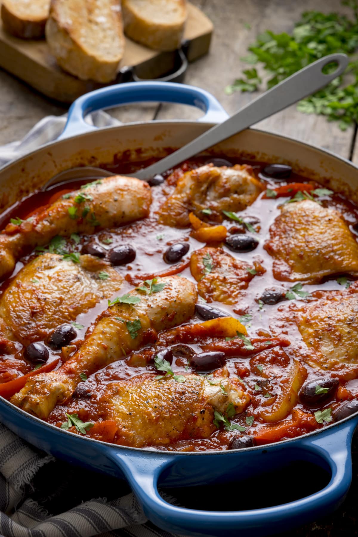 Tall image of chicken cacciatore in a large, shallow blue casserole dish, with a silver serving spoon sticking out. The dish is on a wooden table, next to some fresh herbs and sliced bread.