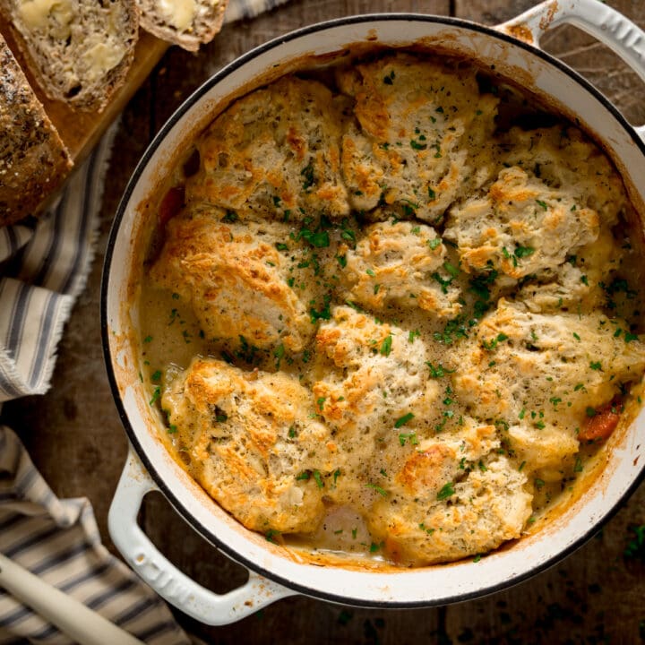 Square image of chicken stew and dumplings in a white casserole pan. The pan is on a wooden table. There is a striped napkin and a loaf of bread with some slices cut and buttered next to the pan.