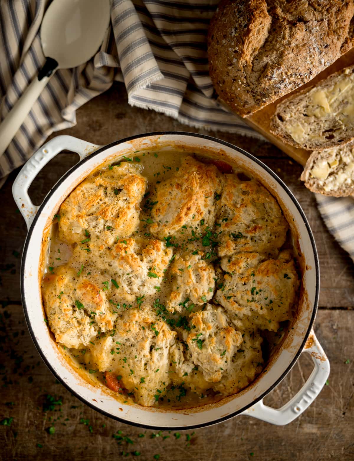 Overhead image of chicken stew and dumplings in a white casserole pan. The pan is on a wooden table. There is a striped napkin and white serving spoon next to the pan. There is also a loaf of bread with some slices cut and buttered next to the pan.